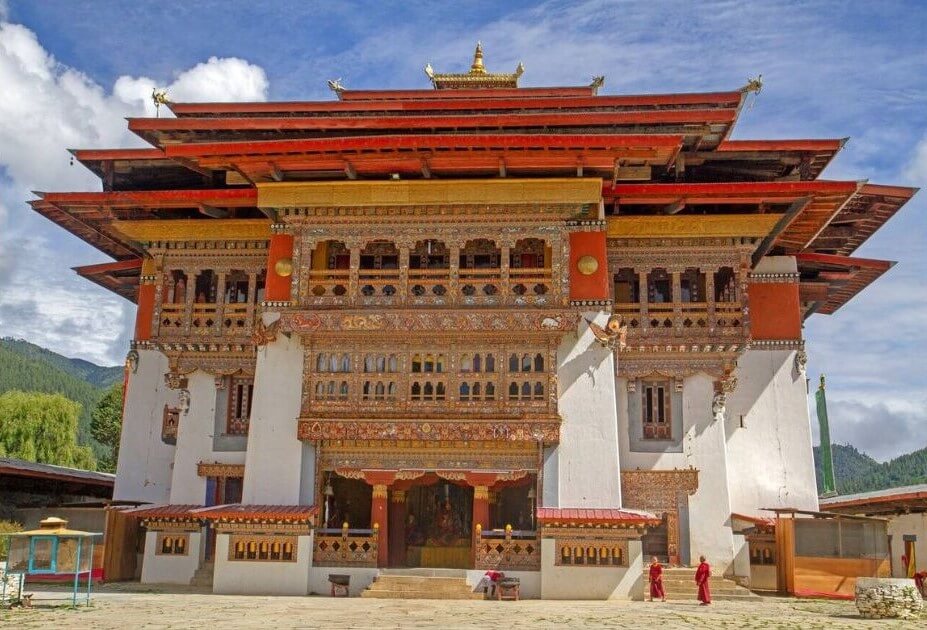 這張圖片展示了不丹著名的崗提寺(Gangtey Goemba),這座傳統不丹建築以其精美的雕刻和鮮明的紅橙色屋頂為特點。寺廟前有兩位僧侶走過,遠處是藍天白雲,背景中還有綠意盎然的樹林,增添了寧靜的氛圍。