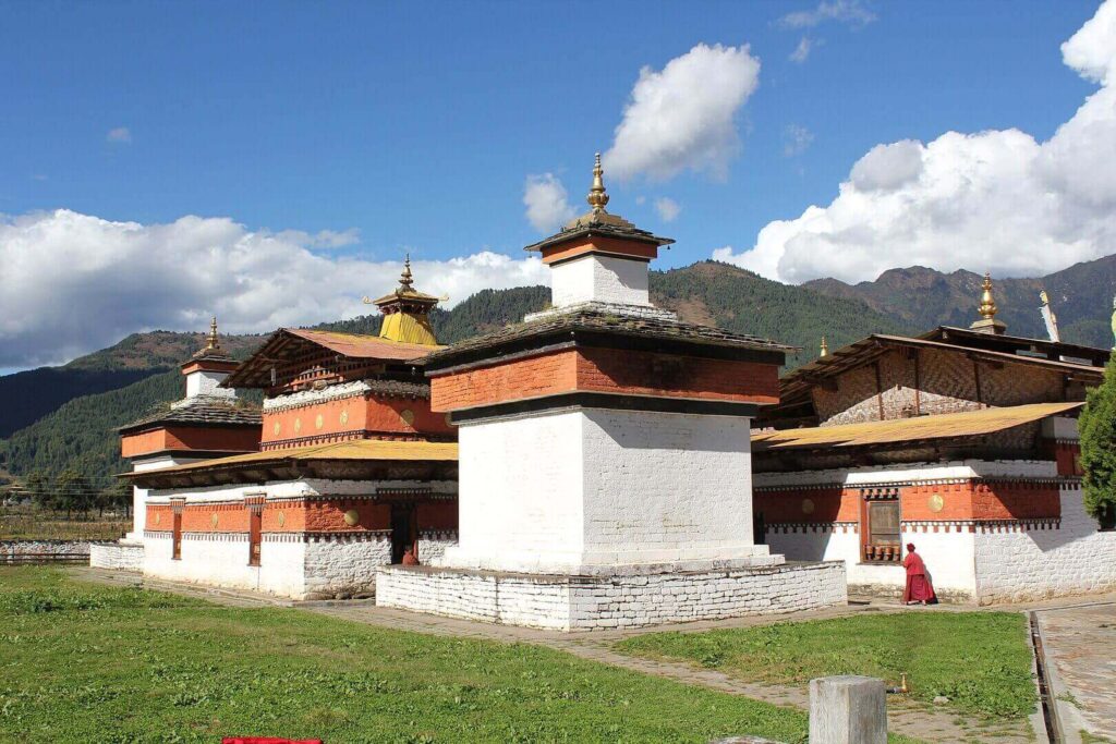 這張圖片展示了不丹的簡培寺(Jambay Lhakhang),這座歷史悠久的佛教寺廟有著紅白色的外牆,周圍被綠地和山脈環繞,背景是一片藍天白雲,畫面中還能看到一位僧人在寺廟旁經過。