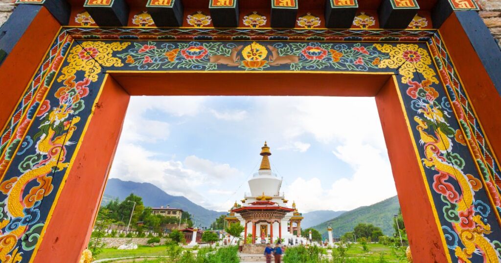 這張圖片展示了國家紀念塔的正門，門框上繪有精美的花紋和龍圖騰。背景中可以看到白色的佛塔和藍天，象徵著不丹人民對和平與繁榮的祈願。