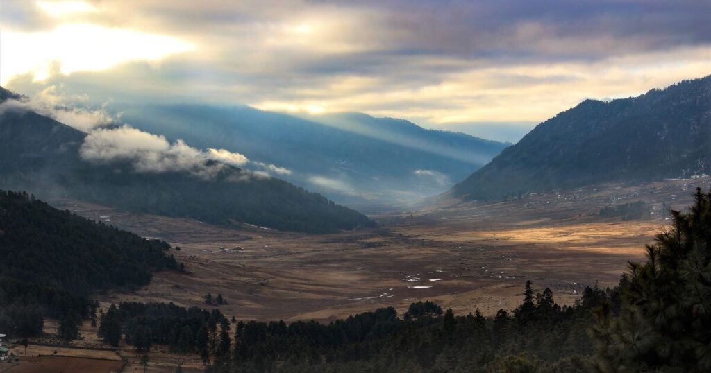 該圖片展示了Phobjikha山谷的壯麗景色，晨光照耀在山谷中，山巒間飄著雲霧，平坦的谷地與遼闊的天空形成了美麗的自然景觀。這是黑頸鶴的重要棲息地，展現了不丹大自然的寧靜與美好。