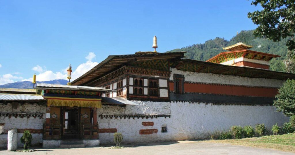 這張圖片展示了不丹的淡心寺(Tamshing Lhakhang),這座歷史悠久的佛教寺廟建築風格獨特,紅白色的外牆與背後的青山相映,給人一種莊嚴和寧靜的感覺。