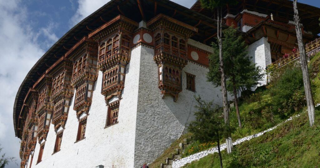 此圖片展示了不丹登古寺(Tango Monastery)的一部分建築,寺廟位於山坡上,白色牆面與精緻的木質窗戶構成鮮明對比。建築風格充滿不丹傳統元素,位於綠樹環繞的自然環境中,彰顯了該寺廟的宗教文化意義與自然和諧的氛圍。