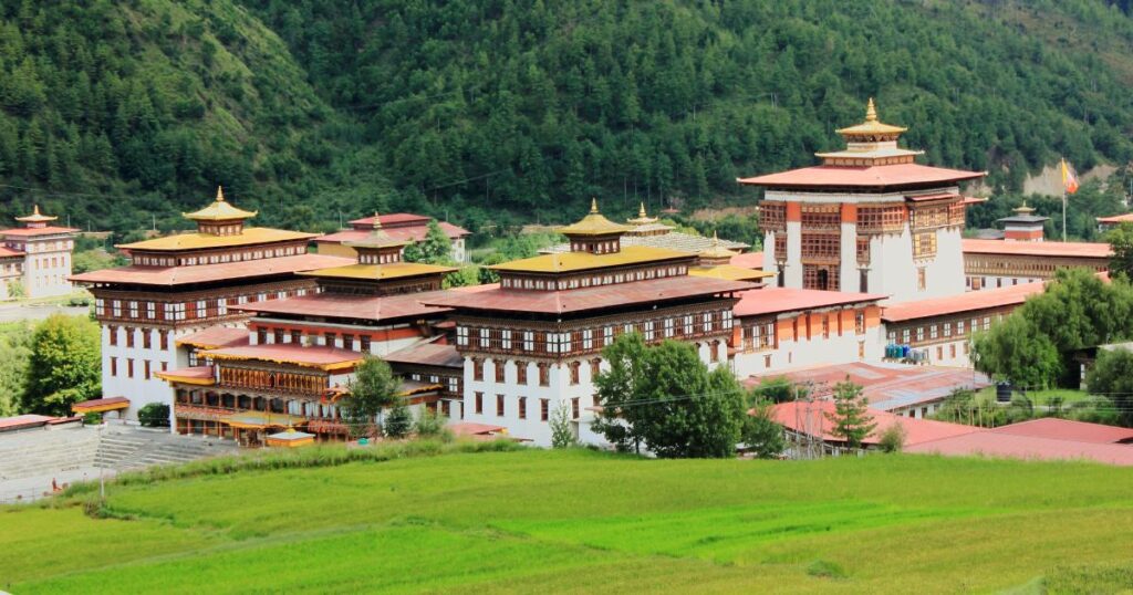 這張圖片展示了位於不丹首都廷布的扎西秋宗，這座傳統宗堡建築以其精美的木雕和白牆紅屋頂聞名。周圍是綠色山丘和廣闊的田野，宮殿式的建築突顯了不丹傳統文化和宗教的重要性。