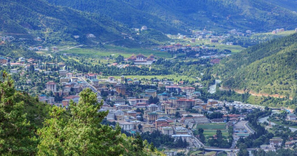 這張圖片展示了不丹首都廷布的壯麗鳥瞰圖，周圍環繞著綠意盎然的山脈。可以清楚看到城市中的寺廟和現代建築融合，展現出不丹獨特的文化與城市景觀。
