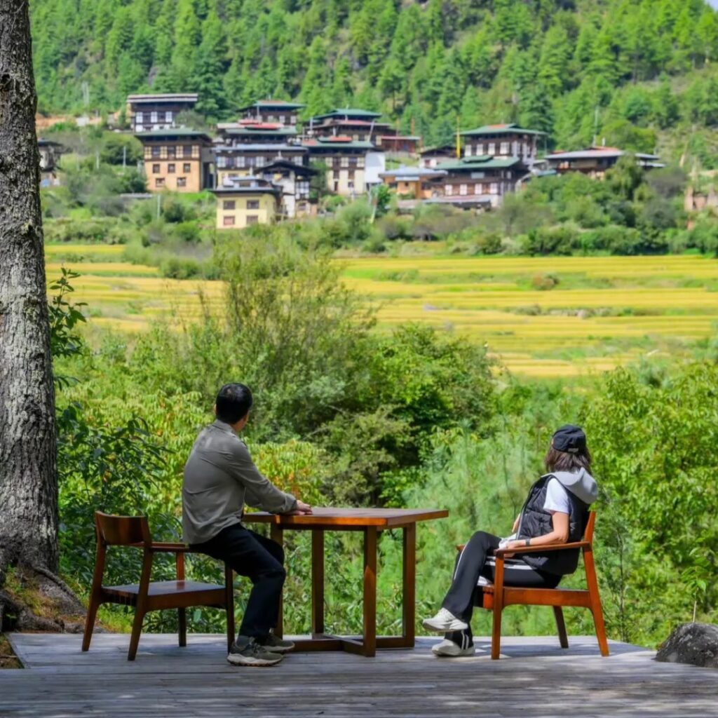 劉嘉玲和梁朝偉坐在戶外木椅上，面向遼闊的綠色山谷與遠處的建築物。背景是充滿層次感的山丘與農田，展示了不丹自然景觀的美麗與寧靜氛圍。