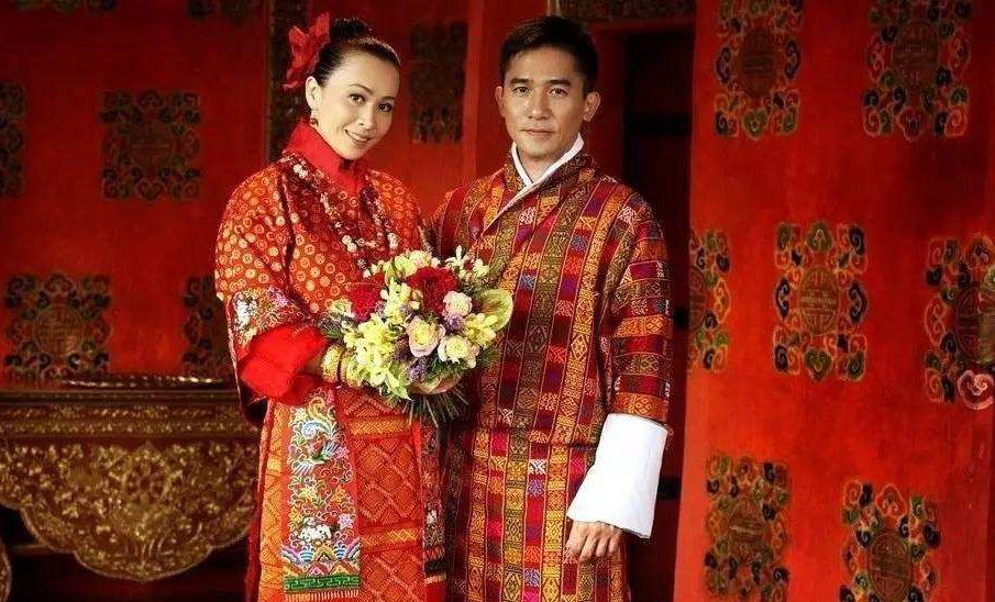 劉嘉玲與梁朝偉身穿傳統不丹服飾，背景是一片鮮紅的牆壁，裝飾有不丹傳統紋樣。兩人手捧鮮花，站在一起，展現出婚禮的莊嚴與喜慶氛圍，彰顯了不丹文化中的細膩與色彩繽紛的藝術風格。