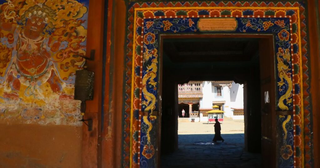 這張圖片展示了一個不丹寺廟的彩繪入口，雕刻精美的彩色圖案裝飾了門框。門內透出一位僧侶的身影，展現出寺廟內部的神秘氛圍。