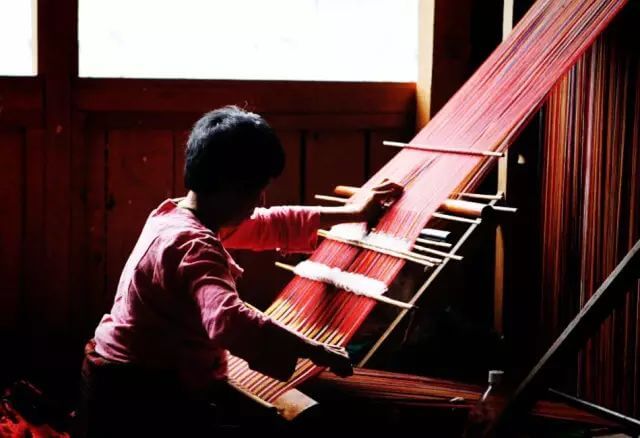 該圖片展示了一位不丹婦女在傳統編織機上編織彩色織物,強調了不丹傳統工藝與文化的重要性,背景中的光影凸顯了編織的專注和細緻。