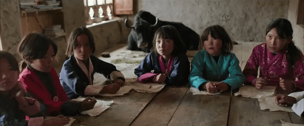 這張圖片展示了一群不丹學生聚集在木製桌旁，專注地望著前方。他們穿著傳統不丹服飾，背景中可見一頭牦牛，環境充滿鄉村氣息，讓人感受到不丹學習環境的純樸自然。