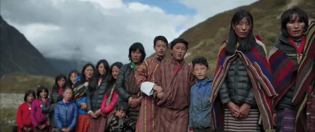 圖片展示了不丹魯納納的村民們在草原上集合，男女老少身著傳統服飾，表情莊重而真誠的歡迎新任國小教師的到來。