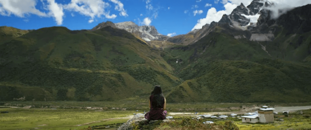 這張圖片展示了一位學生坐在山崖上，凝視著遠方的山谷和壯麗的山脈，背景的雪山與綠意盎然的山坡形成鮮明對比。此場景讓人感受到不丹壯麗的自然景觀與寧靜的氛圍。
