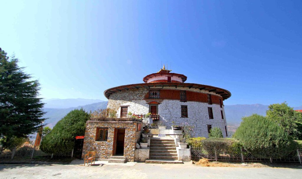 該圖片展示了位於不丹帕羅的國家博物館建築，其圓形的石造結構在藍天映襯下顯得宏偉壯觀。博物館外牆以白色石材和紅磚構成，呈現出傳統不丹建築的特色，屋頂上方裝飾有金色屋頂。周圍綠樹環繞，遠處可以看到連綿的山脈，展現出不丹自然景觀的寧靜與莊嚴。
