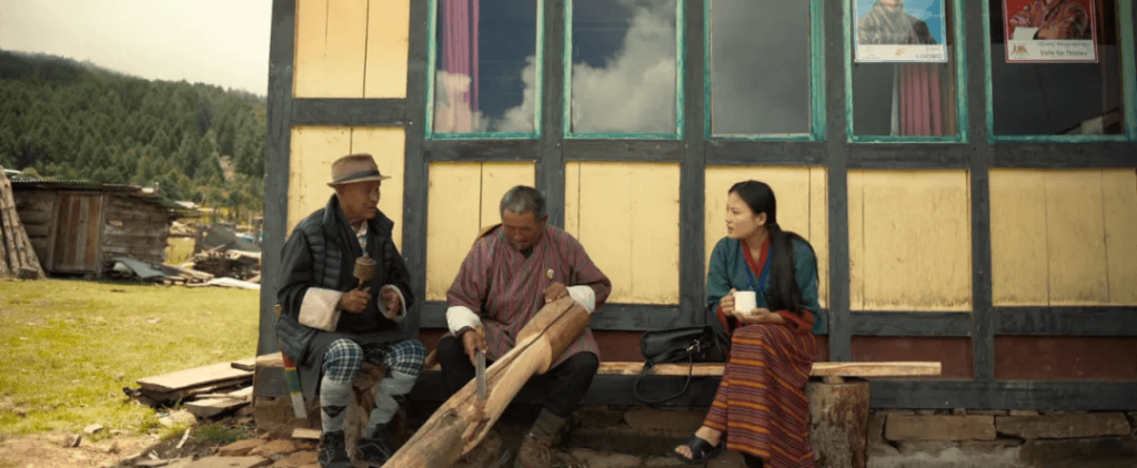 該劇照展示了兩位不丹村民與一位女子在簡樸的建築外談話的場景。村民們手持工具，背景是一間傳統不丹房屋，表現出日常生活中的樸實風貌。