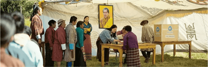 劇照展示了一個社區活動場景，居民們排隊在帳篷內進行登記，背景掛有不丹國王的畫像。此場景展示了不丹人民的社會活動與互動，突顯了村落的凝聚力。