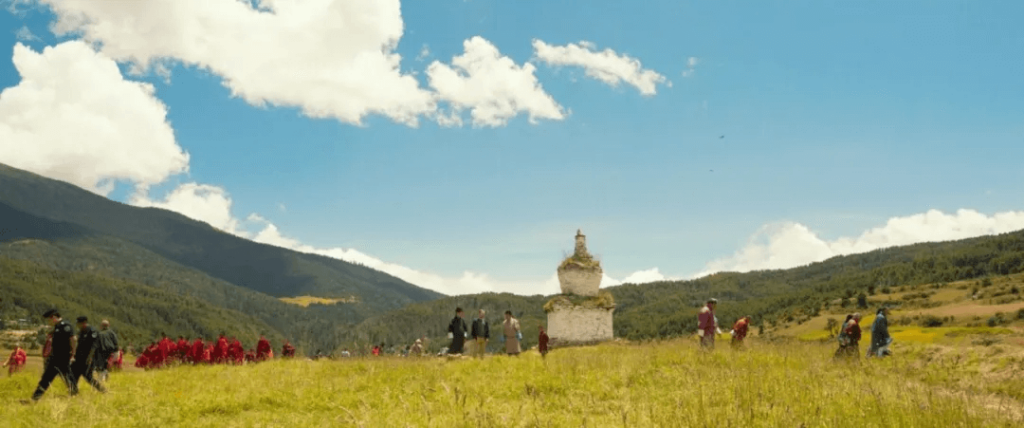 這張劇照捕捉了不丹山區壯麗的景色，演員和劇組成員站在一片開闊的山間草地上。藍天白雲下，遠方的白塔與群山相互映襯，營造出和平祥和的氛圍。