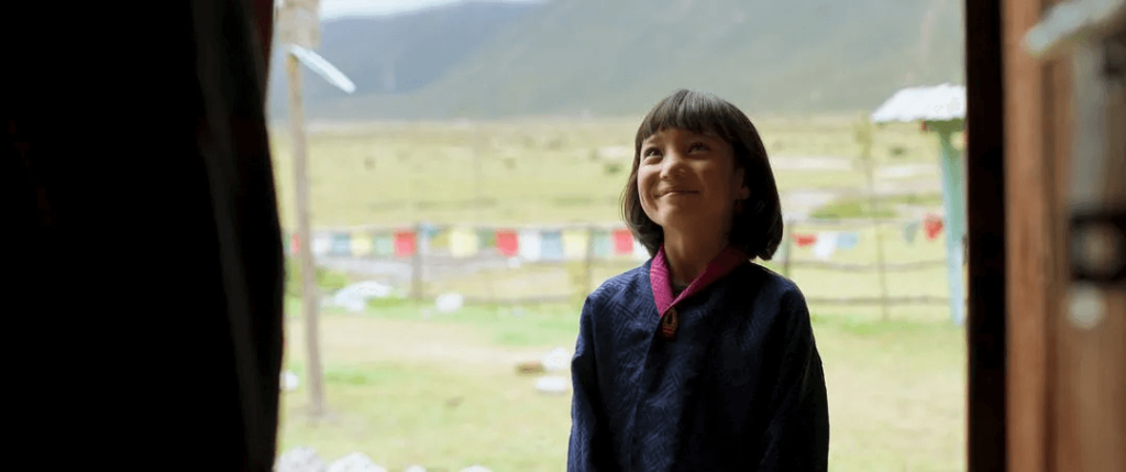 圖片中一位年輕的不丹學生站在門口，露出燦爛的微笑，背景是遼闊的草地與五彩的經幡。她的笑容讓人感受到不丹學生純真的喜悅與平靜的學習環境。