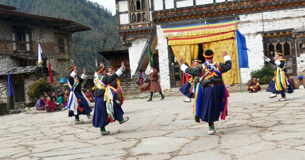 圖中展示了不丹傳統舞蹈表演，舞者穿著色彩鮮艷的民族服飾，在寺廟前方的石板廣場上翩翩起舞，背景是裝飾精美的寺廟建築和青山環繞的美景，氣氛充滿了濃厚的文化色彩與歡樂的節慶氛圍。
