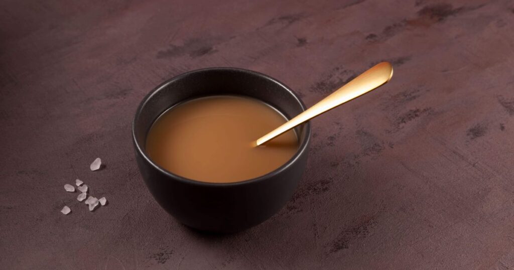 圖片展示了一杯傳統不丹酥油茶（Suja），這是一種將茶葉與酥油和鹽混合製成的飲品。茶呈棕色，杯旁有幾顆粗鹽粒，象徵其獨特的鹹味。背景是深色桌面，營造出溫暖而沉靜的氛圍。